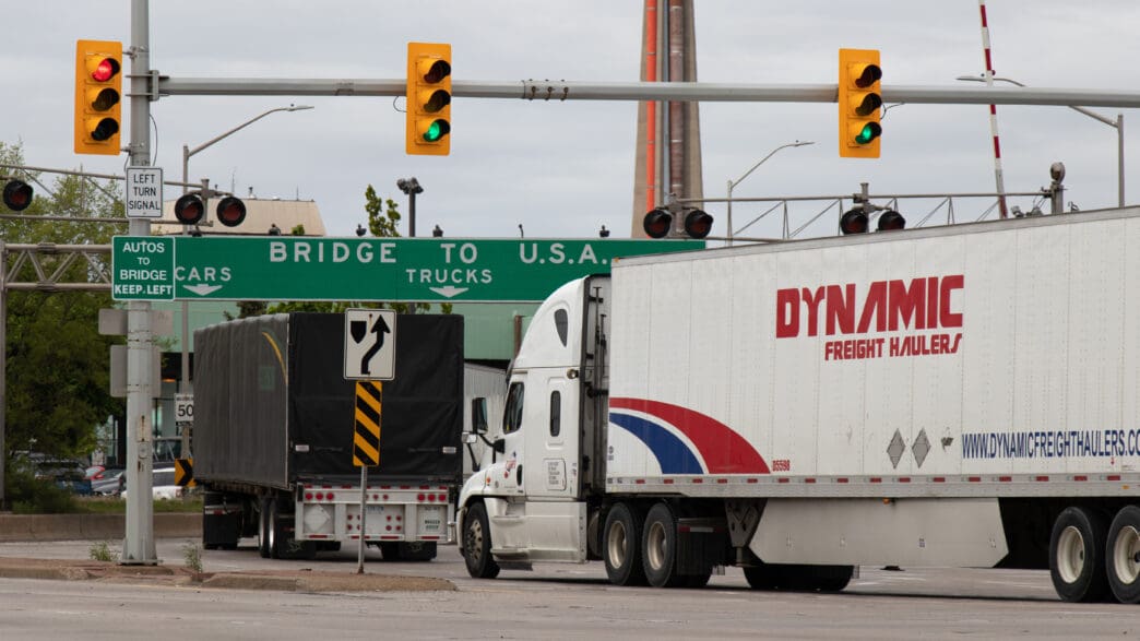 Transport trucks line up under a "Bridge to USA" sign at the US-Canada border crossing.