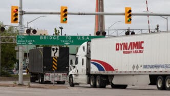 Transport trucks line up under a "Bridge to USA" sign at the US-Canada border crossing.