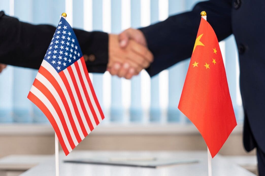 US and China flags in front of diplomats shaking hands during negotiations.