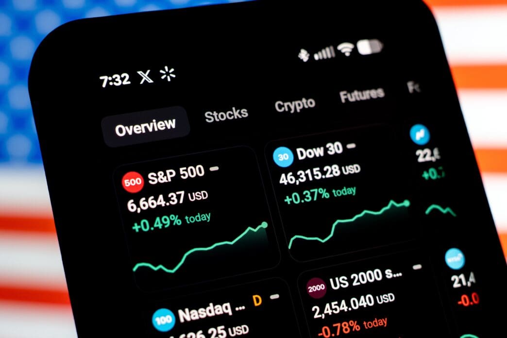 A smartphone screen shows US stock market indices like the S&P 500 and Dow, with a blurred American flag in the background.