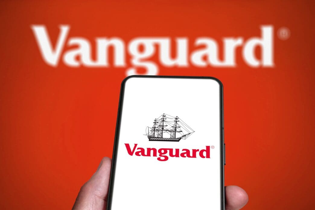 A smartphone displays the Vanguard logo, held in front of a larger, out-of-focus version of the same logo on a red background.