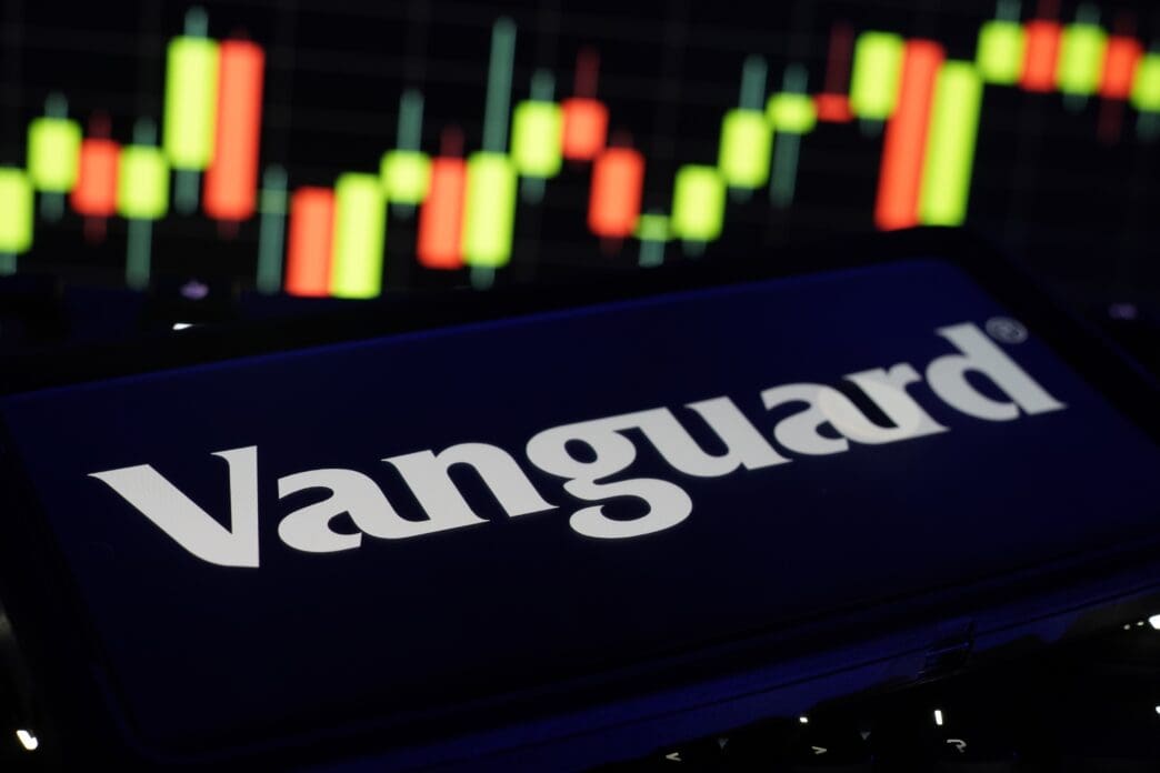 The Vanguard logo on a smartphone screen displayed against a blurred stock market candlestick chart.