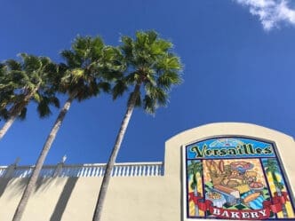 Low angle view of the Versailles Bakery sign and palm trees against a blue sky.