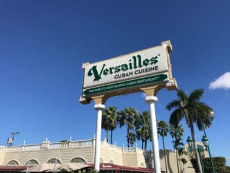 The prominent roadside sign for "Versailles Cuban Cuisine" stands above the restaurant building and palm trees under a clear blue sky.