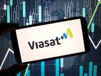 The Viasat company logo is displayed on a smartphone screen, held in front of a blue background with digital financial candlestick charts.