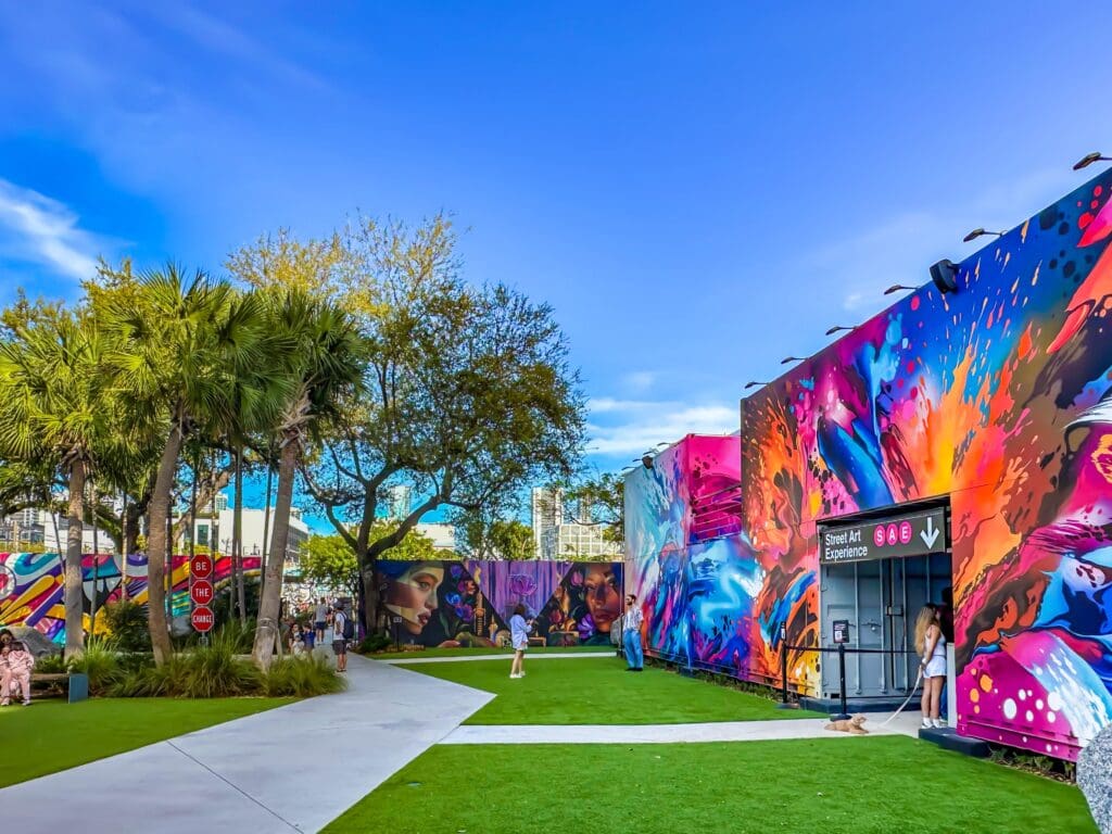 Outdoor view of Wynwood Walls with colorful street murals covering building facades, green lawns, and palm trees under a blue sky.