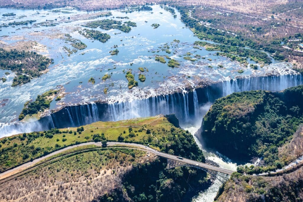 An aerial shot shows the vast Victoria Falls spanning a wide chasm, with the Victoria Falls Bridge crossing the gorge below.