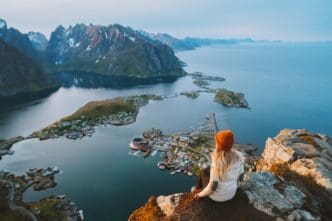A woman wearing a beanie sits on a rocky cliff edge, overlooking the stunning aerial view of the Reine village, fjord, and sharp mountains in the Lofoten Islands, Norway.