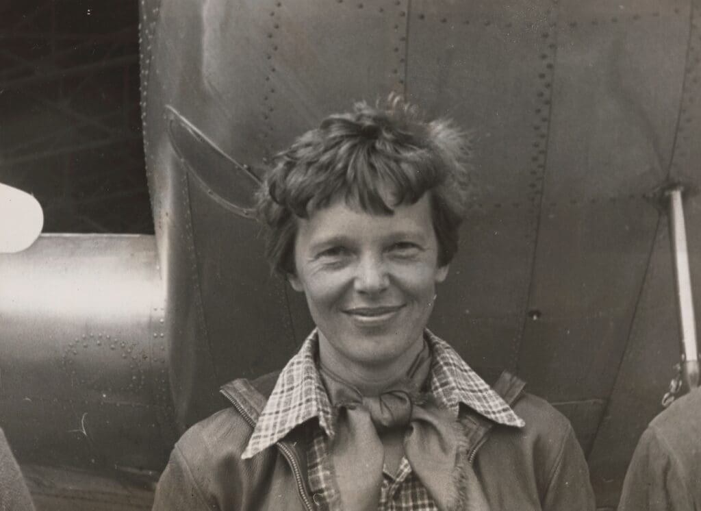 Vintage close-up photo of Amelia Earhart smiling warmly, wearing a leather jacket and checkered shirt with a scarf.