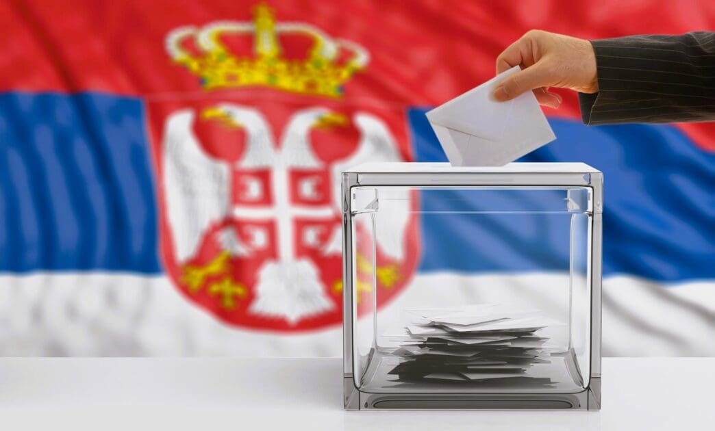 A person's hand deposits a ballot into a clear ballot box in front of a blurred flag of Serbia.