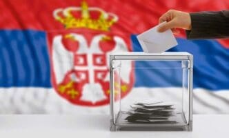 A person's hand deposits a ballot into a clear ballot box in front of a blurred flag of Serbia.
