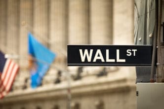 A street sign reading "WALL ST" in white capital letters on a black background, with blurred flags and historic buildings behind it.
