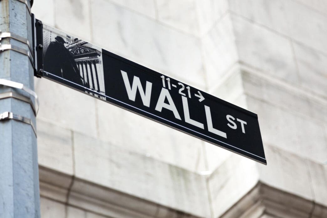A close-up of a black Wall Street street sign attached to a pole, with a marble building facade blurred.