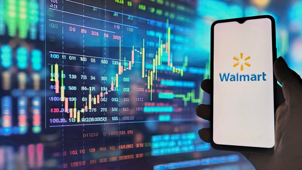 The Walmart logo displayed on a smartphone screen, which is held up against a colorful, out-of-focus digital stock chart.