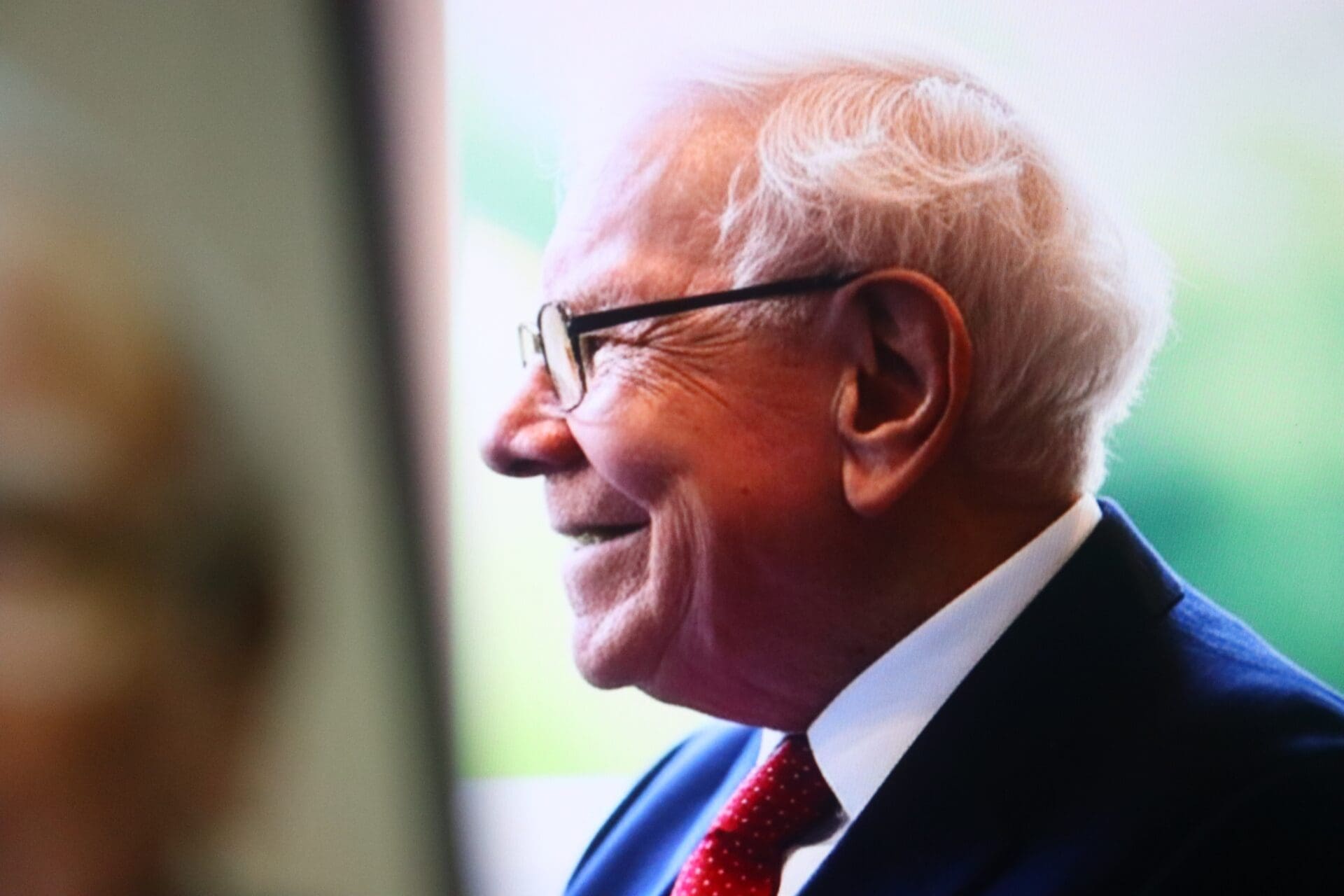 Warren Buffett smiles while wearing glasses, a suit, and a red polka-dot tie.