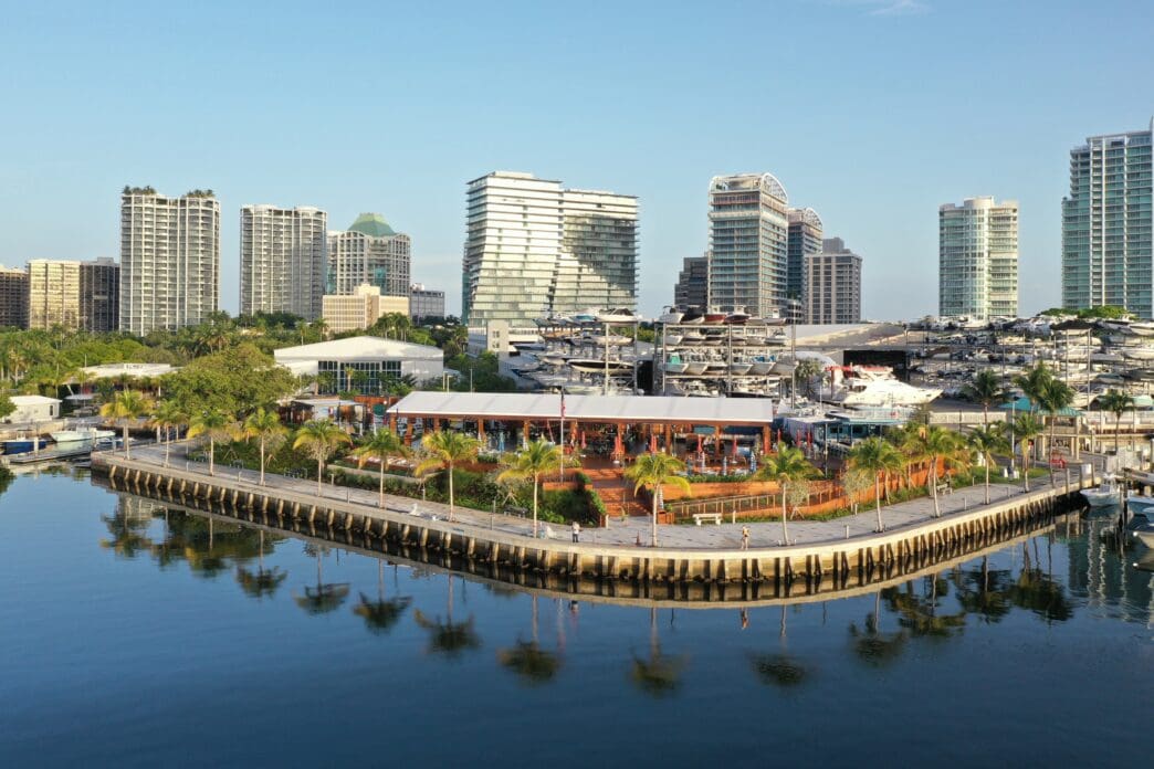 An aerial view of the Coconut Grove waterfront in Miami, showing restaurants, a marina, and the city skyline on a sunny morning.