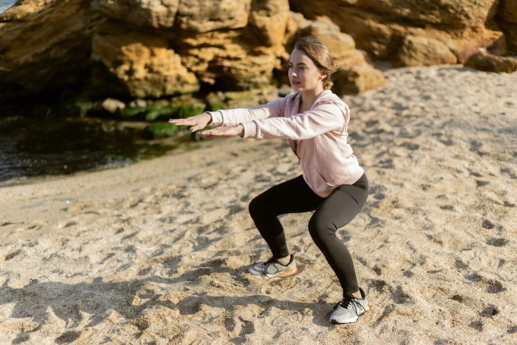 Young woman in a pink hoodie and black leggings performs a squat exercise on a sandy beach.