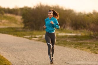 A smiling woman in a blue jacket and grey leggings jogs along a paved path in a park at sunrise.