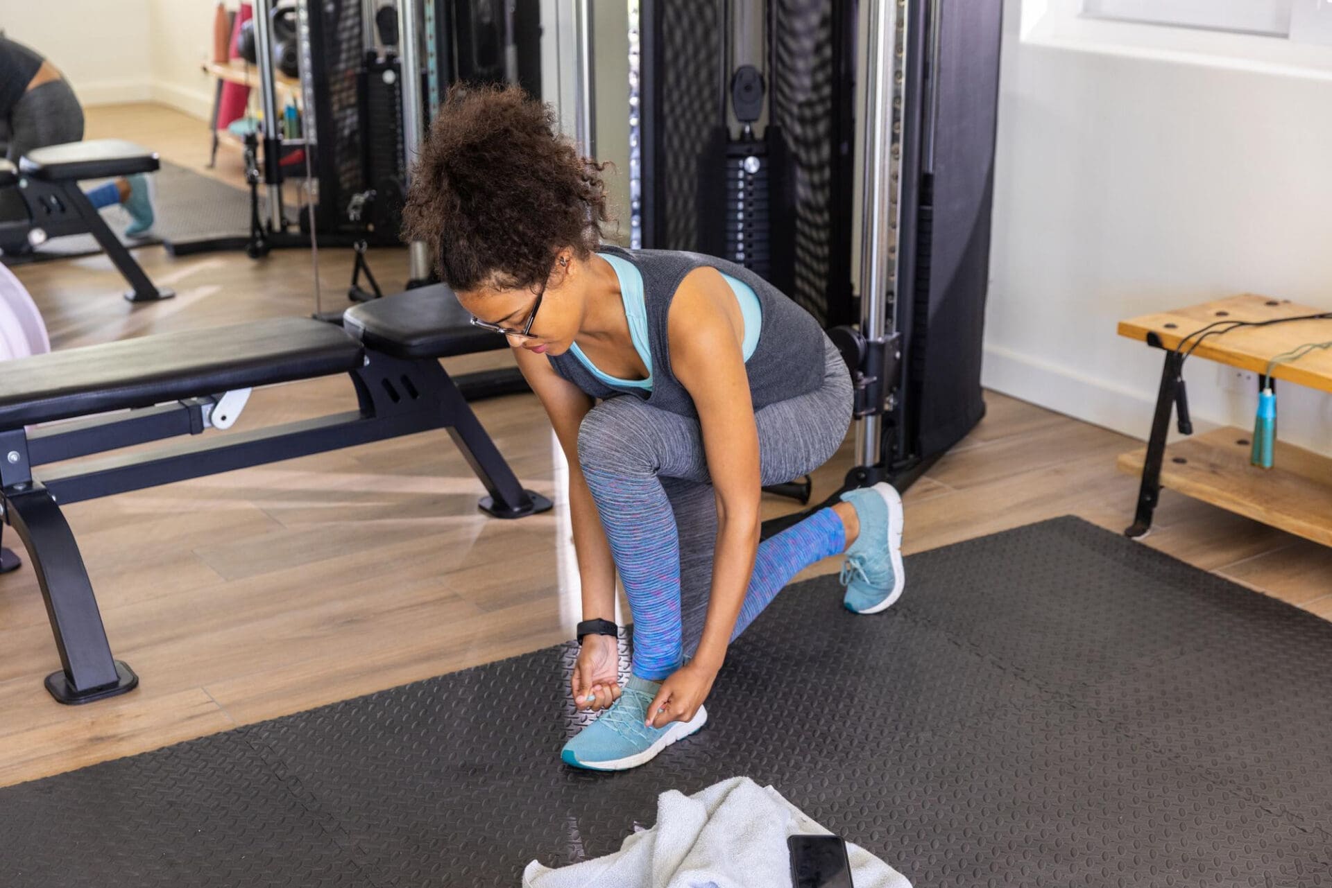 A woman in athletic wear kneels on a black mat in a gym to tie the shoelace of her running shoe.