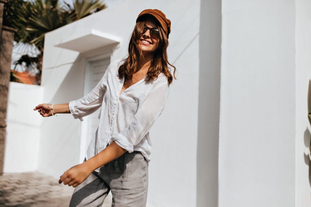 Smiling woman wearing a white linen button-down shirt, a brown cap, and sunglasses stands against a bright white wall outdoors.