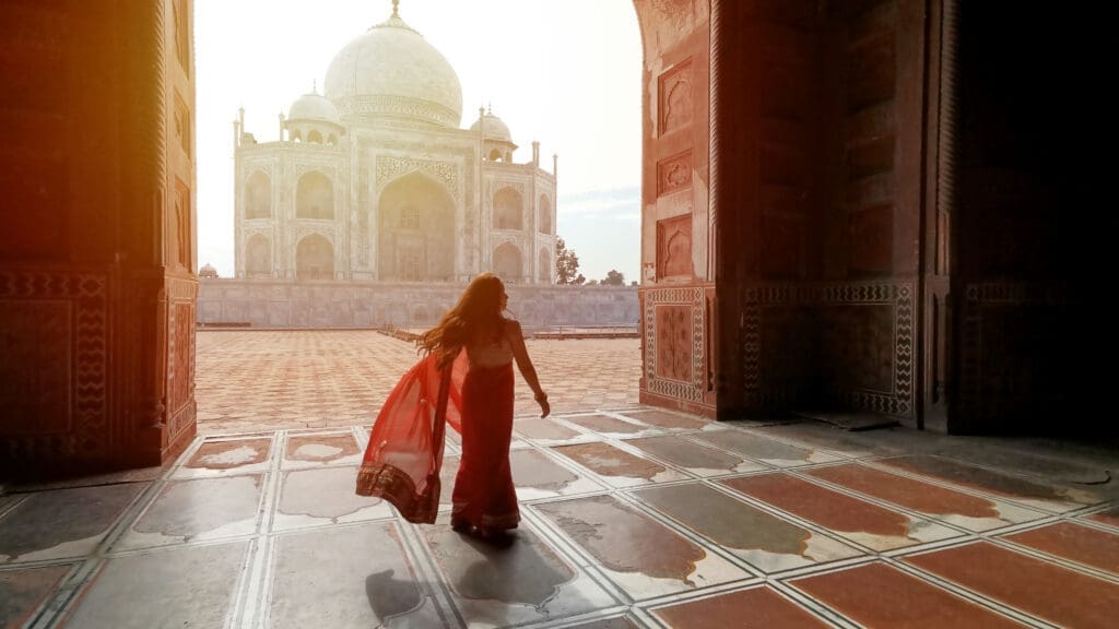 Indian woman in a flowing red sari walks across a stone courtyard toward the sunlit Taj Mahal, framed by a darkened archway.