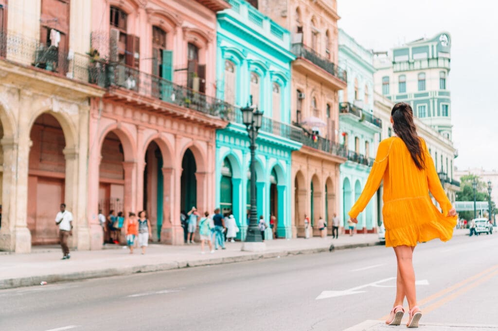 A woman in a bright yellow dress walking down a street past a row of historic pastel-colored buildings in Havana, Cuba.