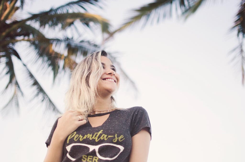 Smiling woman with blonde hair wearing a dark graphic t-shirt featuring stylized text and sunglasses design, viewed under palm fronds.