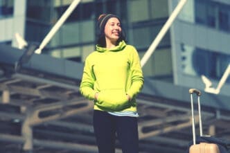 A smiling woman in a green hoodie and beanie stands with a suitcase in a city.