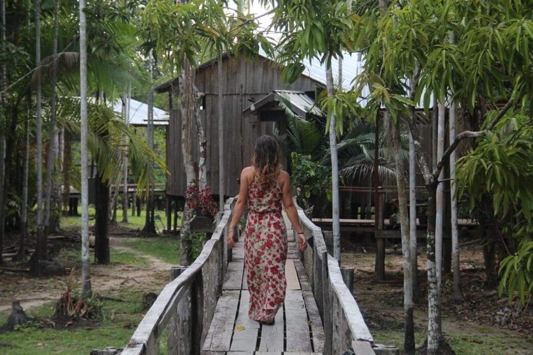 Woman in a long floral dress walks away from the camera on a wooden walkway toward a wooden stilt house deep in a tropical jungle.