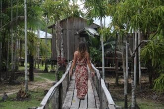 Woman in a long floral dress walks away from the camera on a wooden walkway toward a wooden stilt house deep in a tropical jungle.