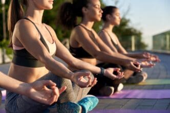 A group of diverse women sit in a row in the lotus pose, meditating outdoors on yoga mats at sunrise.