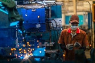 An Asian worker in protective gear stands near a robot auto welding machine, with bright welding sparks visible.