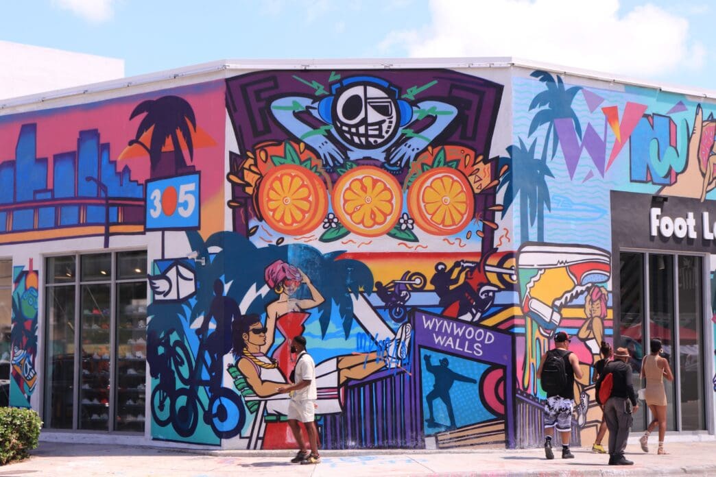 A large, vibrant street art mural covers a corner building in the Wynwood art district of Miami, with pedestrians admiring it.