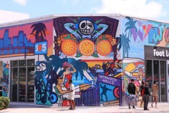 A large, vibrant street art mural covers a corner building in the Wynwood art district of Miami, with pedestrians admiring it.