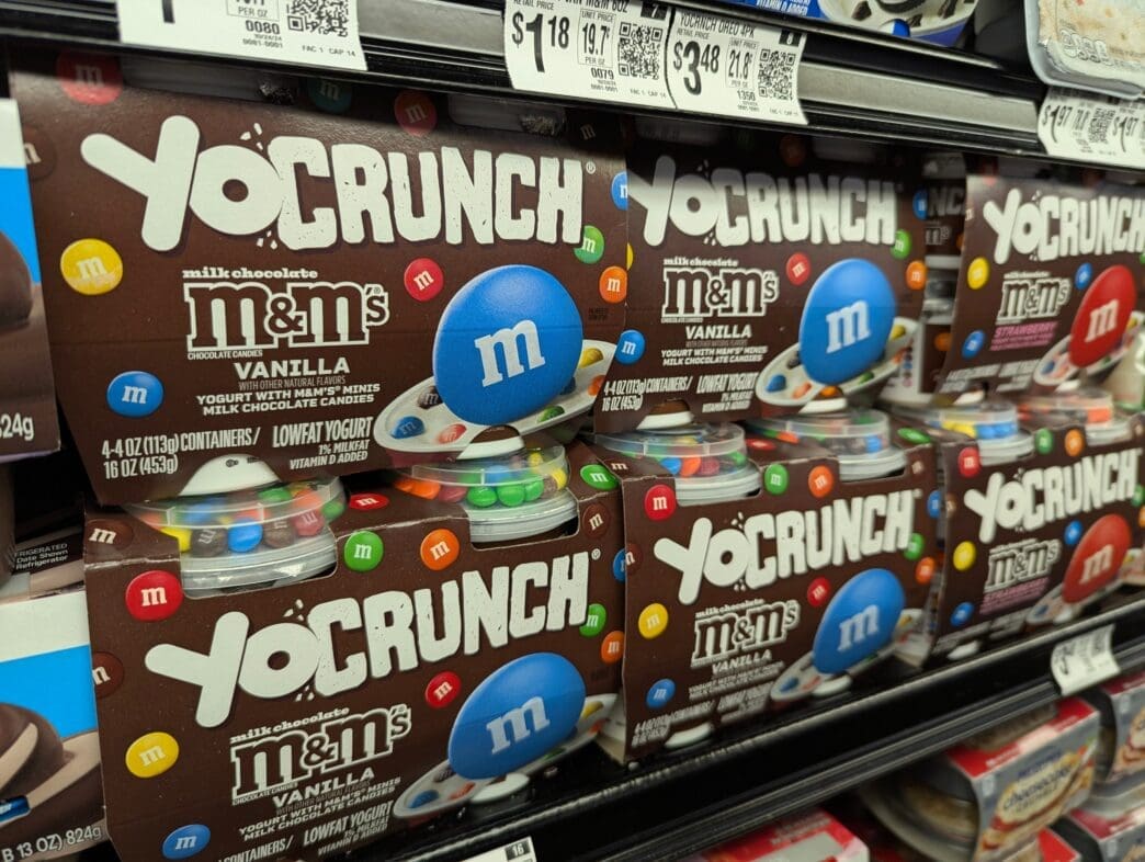 Boxes of YoCrunch Vanilla yogurt with M&M's toppings stacked on a refrigerated shelf at a grocery store