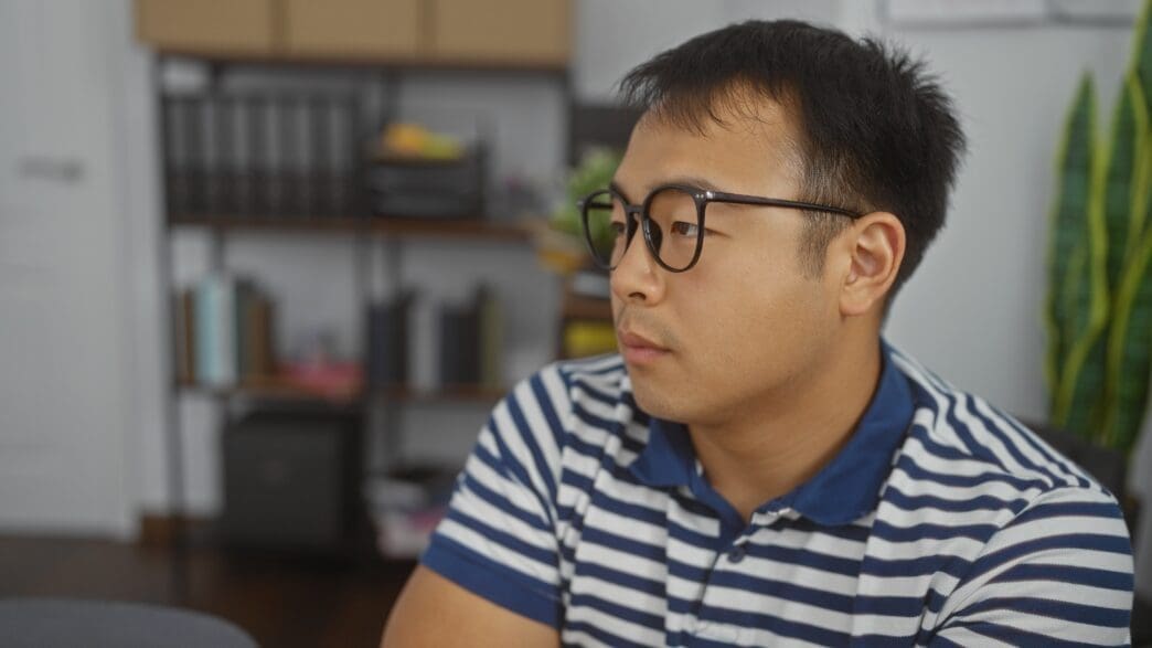 A young Chinese man wearing glasses and a striped polo shirt sits in an office setting, looking off-camera.