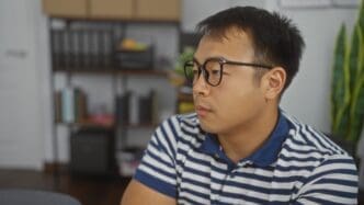 A young Chinese man wearing glasses and a striped polo shirt sits in an office setting, looking off-camera.