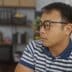 A young Chinese man wearing glasses and a striped polo shirt sits in an office setting, looking off-camera.