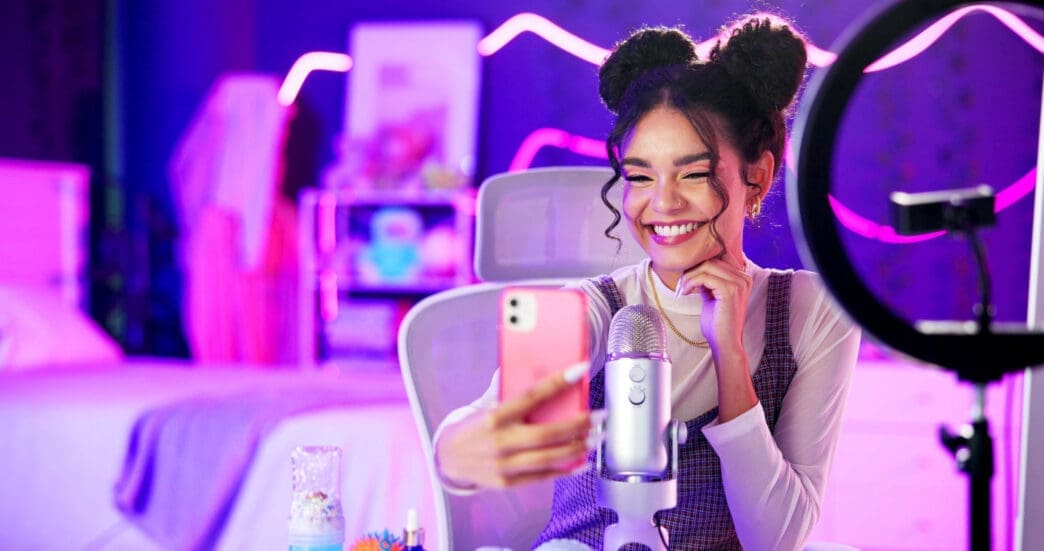 A young, smiling woman with double buns taking a selfie video in a neon-lit pink and purple room with a professional microphone.