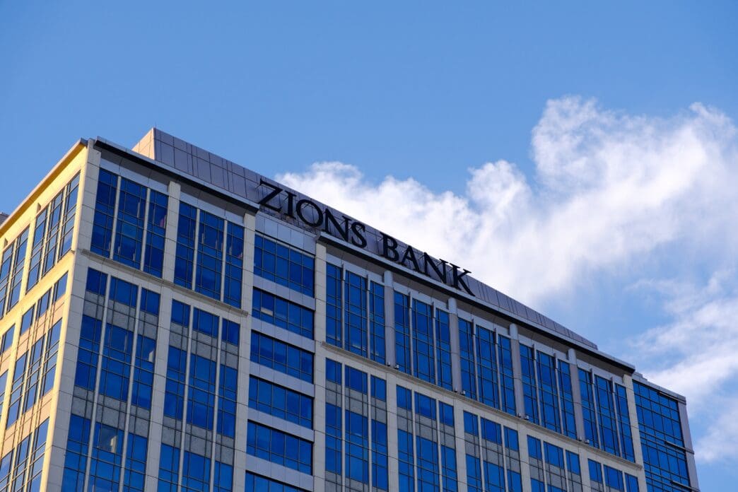 The "ZIONS BANK" name is prominently displayed on the top facade of a modern, multi-story office building with blue reflective glass windows under a bright sky.