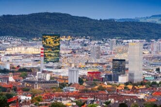 An elevated view of the Zurich, Switzerland skyline at twilight, featuring modern high-rise buildings and traditional red rooftops against a dark, wooded hill.