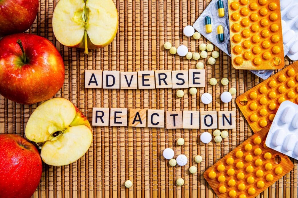 Close-up of apples spelling out the words "Adverse Reaction" next to pills and prescription drugs.