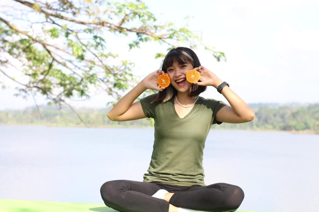 An Asian woman holds two yellow oranges in her hands, lit by morning sunlight.