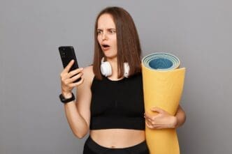 A woman in black sportswear, looking confused and holding a yoga mat, is using her phone against a gray background.