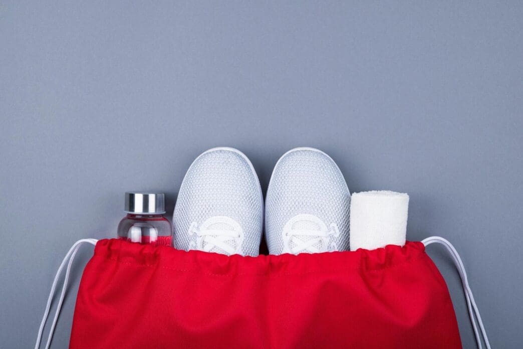 Overhead view of shoes, a towel, and a bottle inside a bag, set against a gray backdrop.