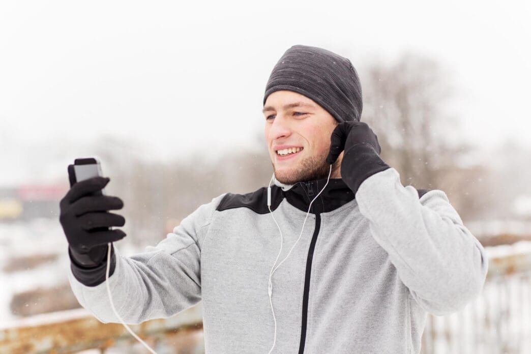 A young man wearing earphones and holding a smartphone smiles while listening to music on a snow-covered bridge.