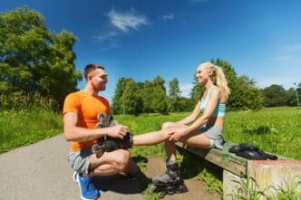 A man helps a woman put on rollerblades outdoors, representing a couple engaging in a healthy summer activity.