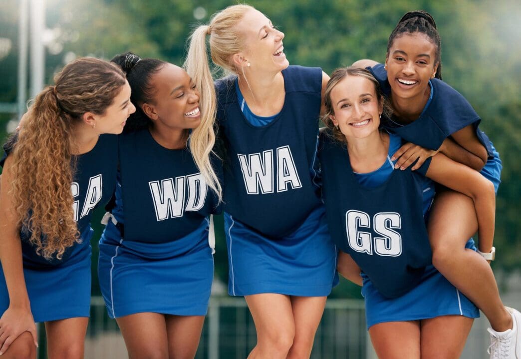 A diverse team of women netball players huddle and hug on a court before a training session.