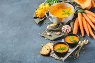 A rustic wooden table displays a still life of autumn ingredients, including a roasted orange pumpkin, carrots, and soup, representing a seasonal diet and nutrition concept.