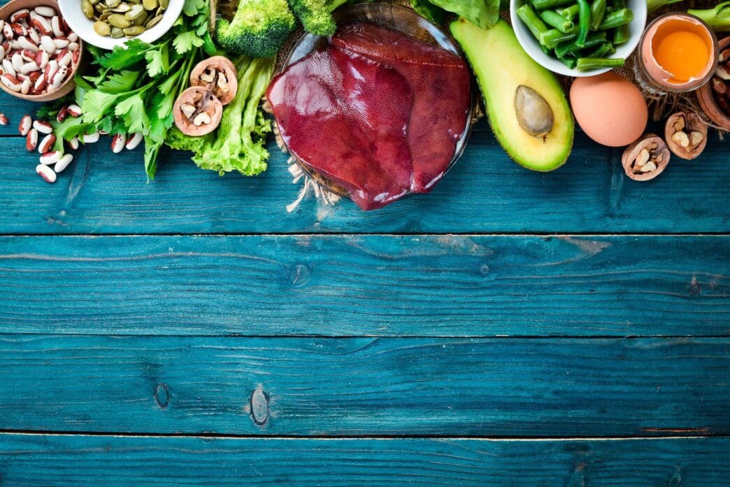 Overhead view of a blue surface with various foods high in vitamin B9, including liver, avocado, broccoli, spinach, parsley, beans, and nuts.